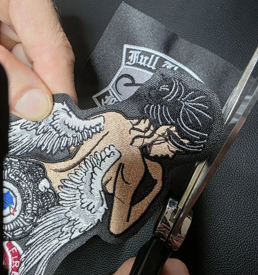 A hand steadying an embroidered patch while scissors trim the last of the border away.