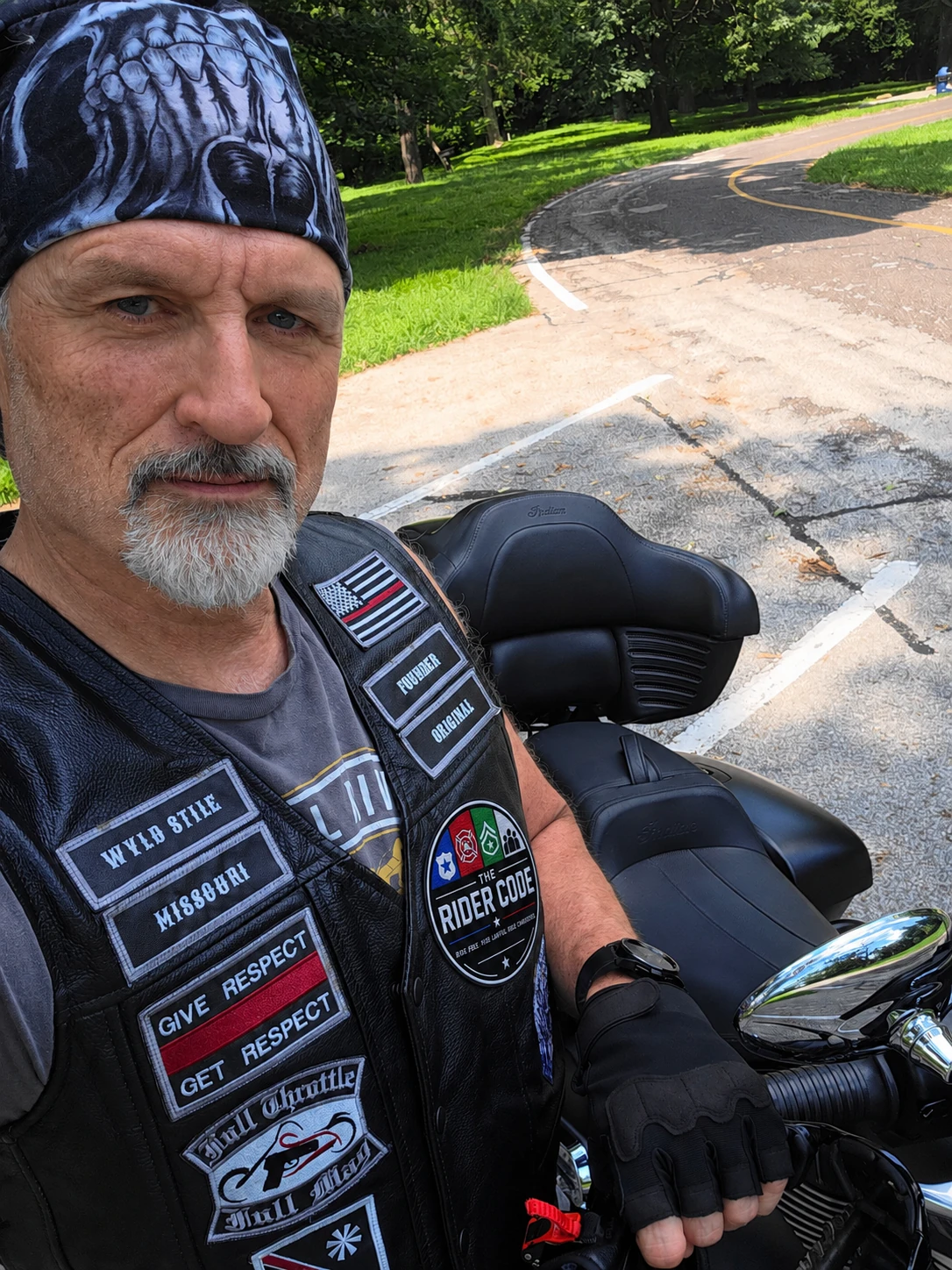 The owner of Independence Embroidery in a black leather vest with embroidered name tabs and front patches, beside his motorcycle.