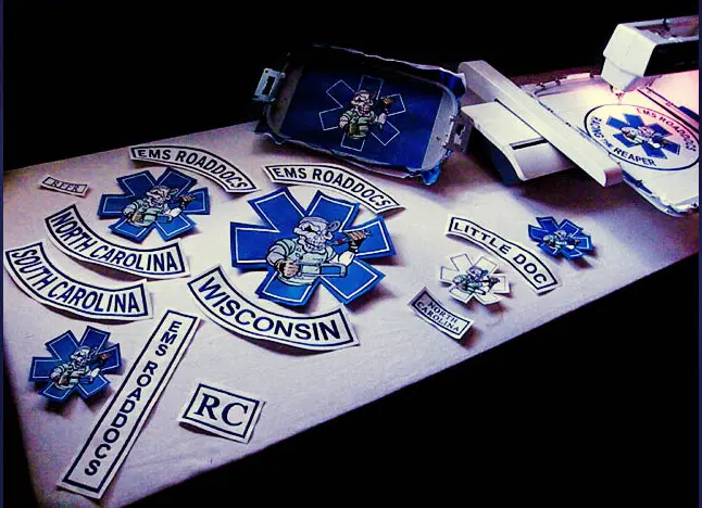 Finished embroidered patches and rockers laid out on a table beside a hooped machine, at Independence Embroidery in Owensville, Missouri.