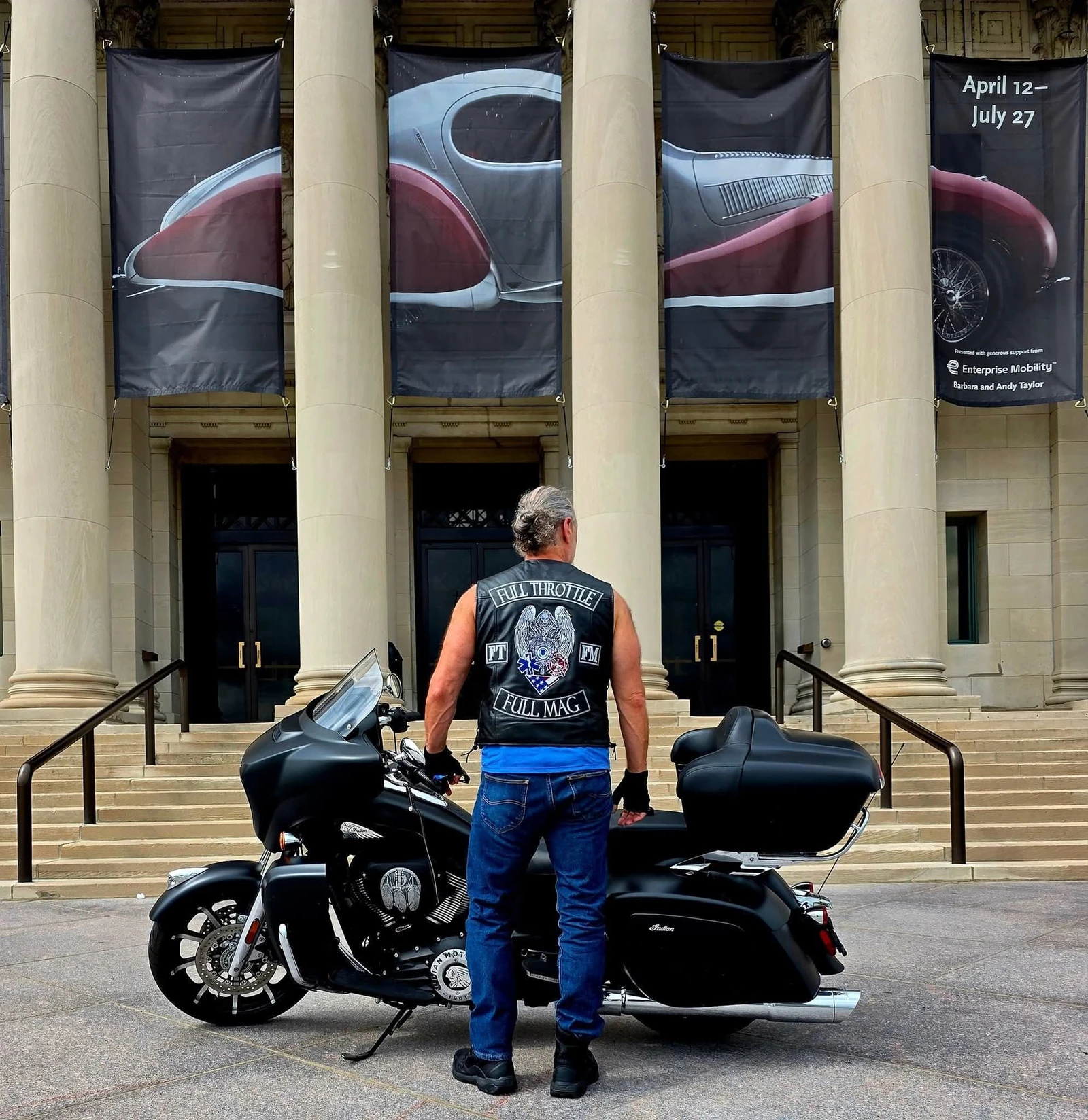 The owner of Independence Embroidery seen from behind in a black leather vest with embroidered Full Throttle Full Mag colors, beside a motorcycle.