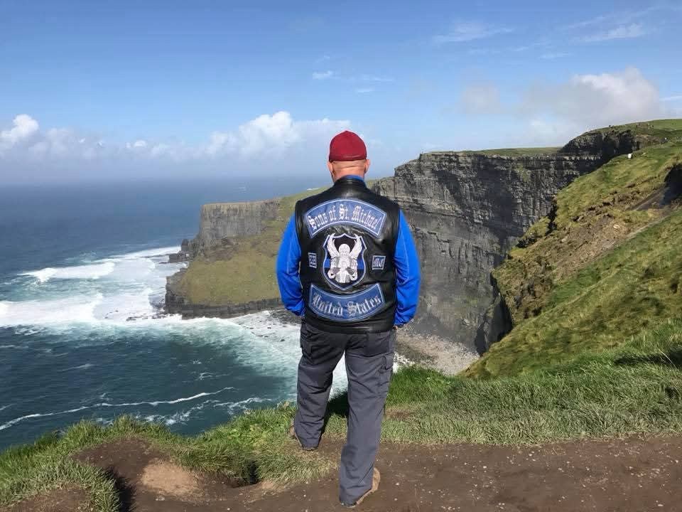 A rider seen from behind in a full set of embroidered club colors, standing on sea cliffs above the surf.