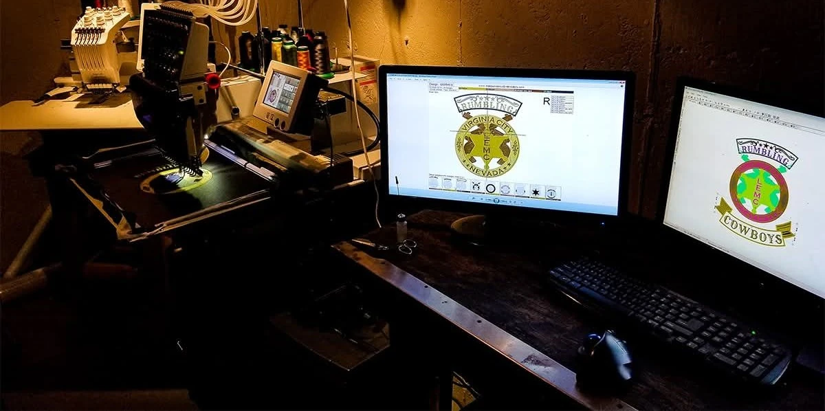 An embroidery machine and two monitors showing club patch designs being digitized on a workbench at Independence Embroidery in Owensville, Missouri.