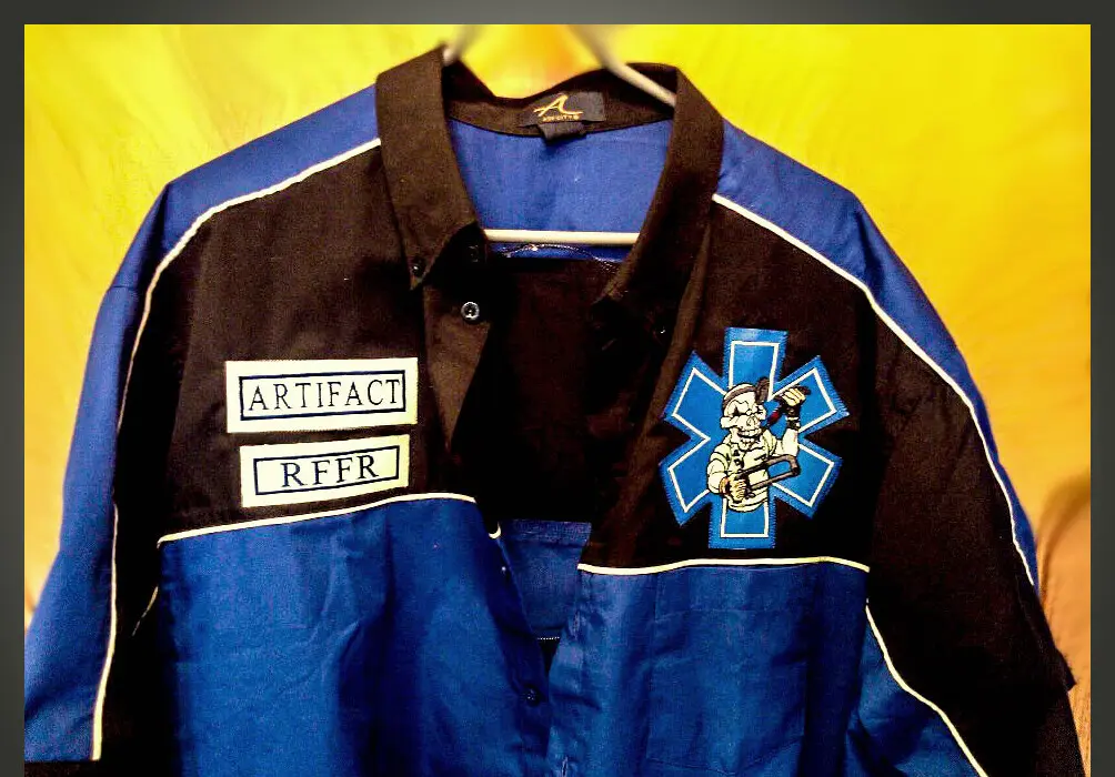 A two-tone blue and black work shirt with an embroidered chest emblem and two name tabs, made by Independence Embroidery in Owensville, Missouri.