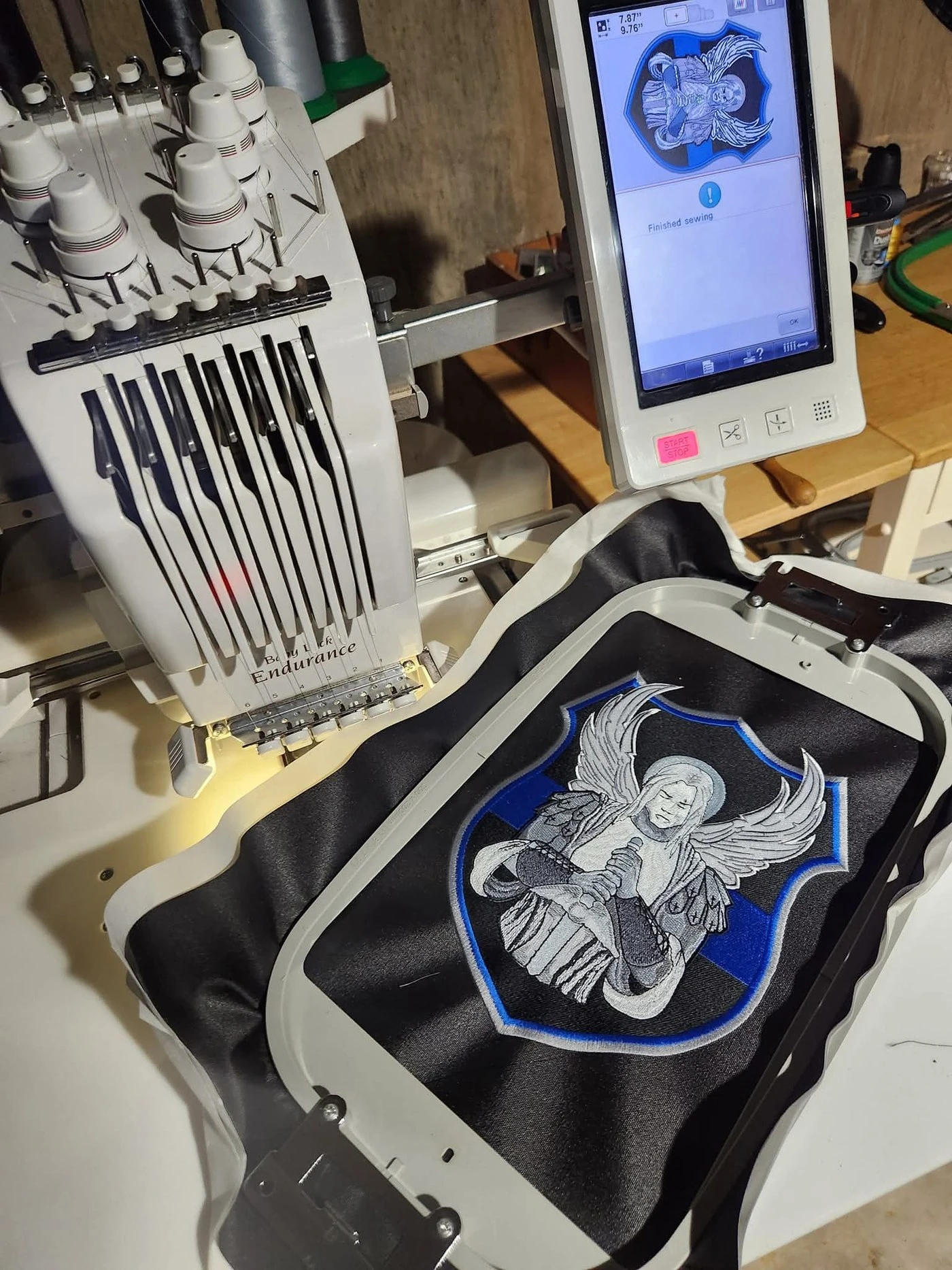 A large embroidered shield patch held in a hoop on a commercial embroidery machine, the machine screen showing the run finished, at Independence Embroidery in Owensville, Missouri.