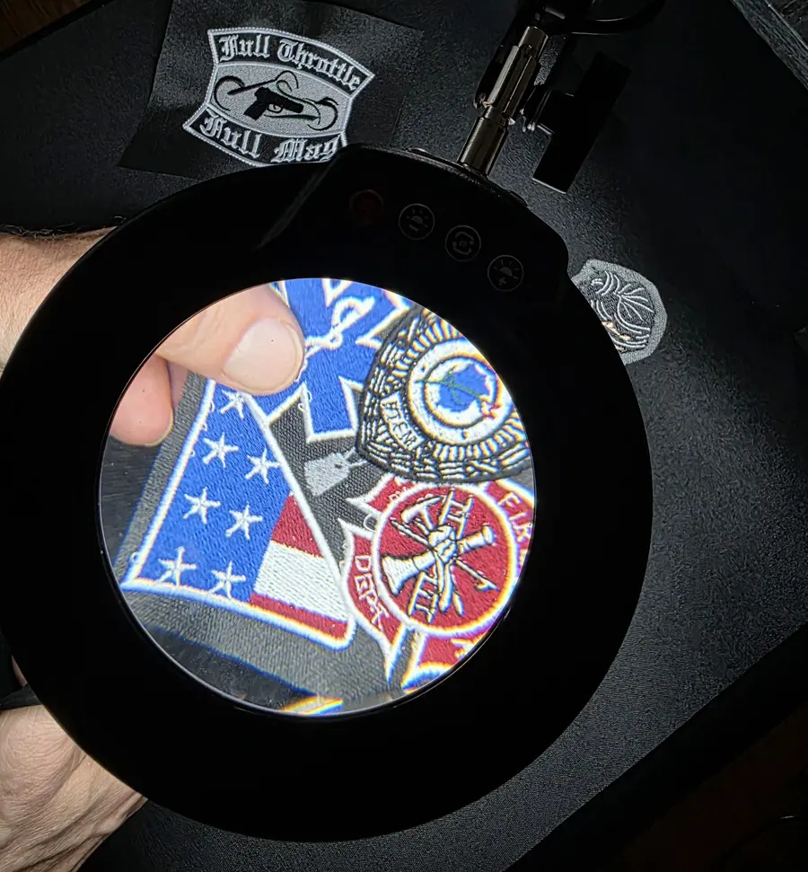 A finished embroidered patch inspected under a lit magnifier at Independence Embroidery in Owensville, Missouri.