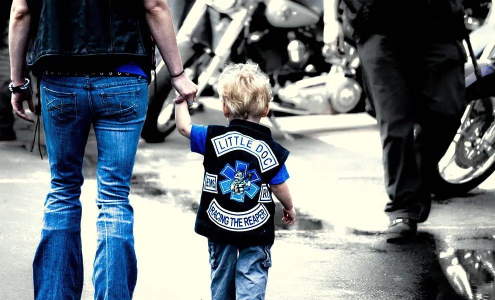 A small child in a miniature embroidered vest with a top rocker, center patch, and bottom rocker, walking beside an adult, made by Independence Embroidery in Owensville, Missouri.